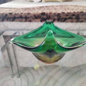 May include: A green glass decorative bowl with a unique, angular shape. The bowl has a pointed center and flared edges, with a dark green and yellow color gradient. The bowl is sitting on a glass table.