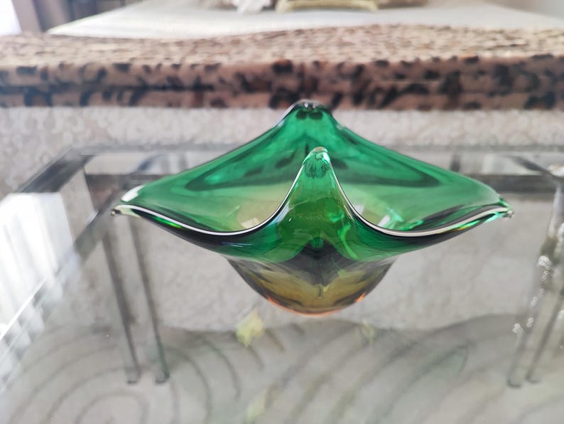 May include: A green glass decorative bowl with a unique, angular shape. The bowl has a pointed center and flared edges, with a dark green and yellow color gradient. The bowl is sitting on a glass table.