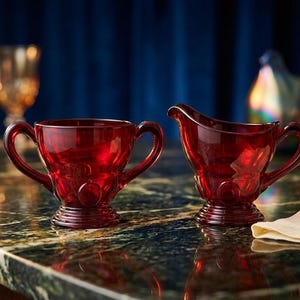 May include: A ruby red glass creamer jug and sugar bowl sit on a marble surface. The creamer has a spout and handle, while the sugar bowl has two handles. Both pieces feature a decorative design.