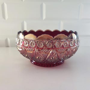 Puede incluir: Un cuenco de cristal rojo rubí antiguo con un borde festoneado y intrincados diseños geométricos. El cuenco se asienta sobre una pequeña base de pedestal y tiene una superficie texturizada detallada. El cuenco es probablemente para servir o exhibir.