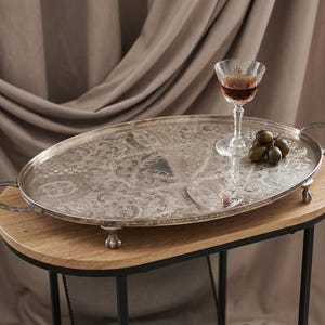 May include: An oval silver serving tray with ornate floral patterns, handles, and small feet. A glass of amber liquid and a cluster of green grapes rest on the tray, which sits on a wooden table. The table has black metal legs.