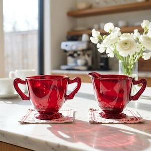 May include: A pair of deep red glass vessels, consisting of a sugar bowl with two side handles and a matching creamer pitcher with a single handle and a pouring spout. Both items feature a pedestal base and a translucent, vibrant ruby finish.