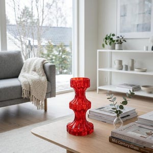 Op de afbeelding: Een levendige rode glazen vaas met een gelaagd ontwerp staat op een lichte houten salontafel. De vaas is het middelpunt, met een grijze bank en een raam op de achtergrond. Boeken en een kleine glazen vaas met groen staan ook op de tafel.