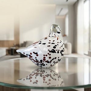 May include: A clear glass bird figurine featuring a rounded body with scattered black and dark red spots throughout the surface. The glass sculpture has a smooth, polished finish and is positioned to show its profile, reflecting on the surface.