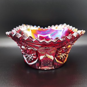 Può includere: Una ciotola di vetro rosso rubino, iridescente, con bordo a balze. La ciotola presenta un motivo geometrico dettagliato all'esterno e un interno vibrante e multicolore. La ciotola è probabilmente vintage o antica.