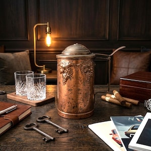 Könnte beinhalten: Ein kupferfarbener Getränkeserver mit Weinreben-Details, zwei Kristallgläser, Zigarren und ein Humidor aus Holz sind auf einem dunklen Holztisch angeordnet. Eine Lampe im Vintage-Stil und ein ledergebundenes Notizbuch sind ebenfalls vorhanden.