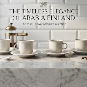 Peut inclure: Quatre tasses et soucoupes à café en céramique blanc cassé de la collection Arctica d'Arabia Finland sont disposées sur un comptoir en marbre. Une cuillère, des grains de café et une cafetière à piston sont également présents. Le texte dit "THE TIMELESS ELEGANCE OF ARABIA FINLAND".