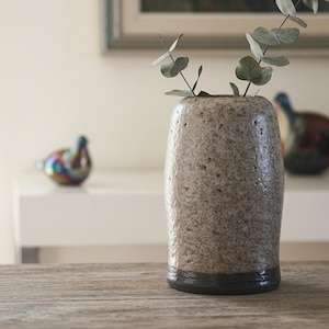 May include: A textured, speckled ceramic vase with a dark rim holds eucalyptus branches. The vase is the focal point, set on a wooden surface. Decorative iridescent bird figurines are in the background.