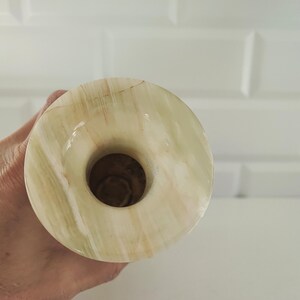 Pu&ograve; includere: Un vaso rotondo in onice verde chiaro e beige con un'apertura centrale. Il vaso ha una superficie lucida e un motivo venato naturale. Lo sfondo &egrave; un muro piastrellato bianco.
