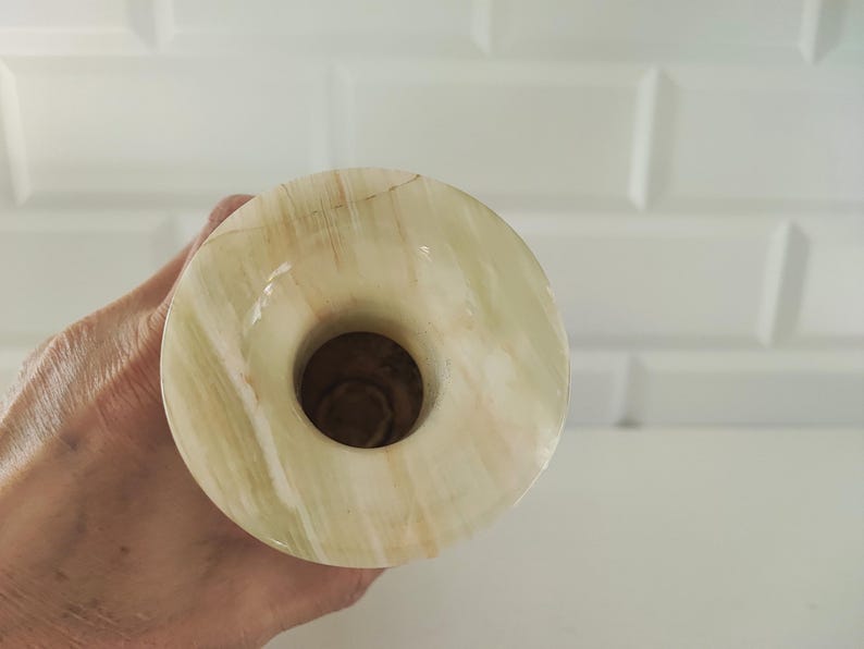 Pu&ograve; includere: Un vaso rotondo in onice verde chiaro e beige con un'apertura centrale. Il vaso ha una superficie lucida e un motivo venato naturale. Lo sfondo &egrave; un muro piastrellato bianco.