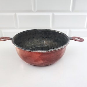 May include: A round copper bowl with a dark, weathered interior and two flat, circular handles extending from opposite sides. The exterior surface shows a warm, metallic copper tone with signs of age, while the rim and interior appear worn.