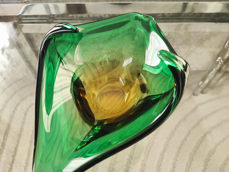 May include: A handcrafted glass bowl with a unique shape, featuring a vibrant green exterior and a golden-yellow interior. The bowl's design is organic and flowing, with a glossy finish. This decorative piece is ideal for home decor.
