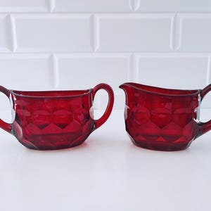 May include: A pair of ruby red glass vessels featuring a faceted geometric pattern. One vessel is a sugar bowl with two side handles, and the other is a creamer pitcher with a single handle and a pouring spout, both made of translucent red glass.