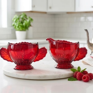 May include: A pair of vintage ruby red glass serving pieces consisting of a sugar bowl with two side handles and a creamer pitcher with a pouring spout and one side handle. Both items feature a scalloped rim and a pressed fan pattern base.