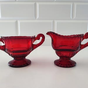 May include: A pair of ruby red glass vessels consisting of a sugar bowl with two side handles and a creamer jug with a single handle and a pouring spout. Both pieces feature a scalloped rim, a ribbed base, and a circular pedestal foot.
