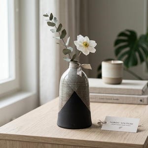 Pode incluir: Um vaso de cerâmica com um design geométrico cinza e preto contém uma flor branca e um galho de eucalipto. O vaso é amarrado com uma fita bege. Uma etiqueta que diz "NORDIC SOLITUDE" repousa sobre uma mesa de madeira.