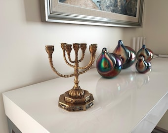 Puede incluir: Un candelabro de metal color latón con cinco portavelas, que presenta un tallo central y una base hexagonal escalonada con grabados decorativos. A su lado hay tres adornos de vidrio iridiscente en forma de lágrima con tonos metálicos oscuros.