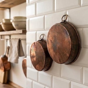 Op de afbeelding: Twee ronde koperen pannen met metalen ophangringen aan de bovenranden. De pannen hebben een verweerde, donkerbruine patina en zijn bevestigd aan een witte, rechthoekig betegelde muur in een keukenomgeving.