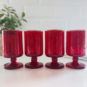 Puede incluir: Un juego de cuatro copas de cristal rojo rubí a juego, cada una con un cuenco cilíndrico, un tallo corto y una base circular. La cristalería tiene un acabado liso y translúcido con un color rojo intenso y uniforme en toda la estructura.