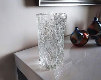 May include: A clear, textured glass vase with a rectangular shape. The vase has a rough, ice-like appearance. Decorative glass spheres with blue, brown, and orange hues are in the background. Home decor.