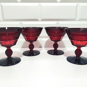 Può includere: Quattro calici da champagne vintage rosso rubino. Ogni calice presenta una fascia strutturata attorno alla coppa, uno stelo rotondo e una base circolare. I calici sono disposti in fila su sfondo bianco.