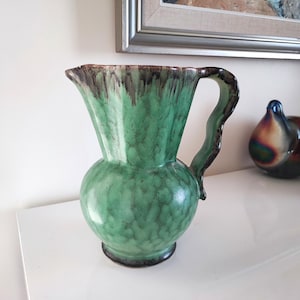 Peut inclure: Une cruche en céramique verte avec une base bombée et un dessus évasé. La cruche a une poignée et une surface texturée. Le bord et la poignée sont brun foncé. Des objets décoratifs sont en arrière-plan.