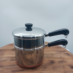 May include: A stainless steel double boiler with a polished finish, featuring two stacked pots and a matching lid. Each pot has a black heat-resistant handle with a small metal hanging loop at the end. The base of the bottom pot is copper-toned.