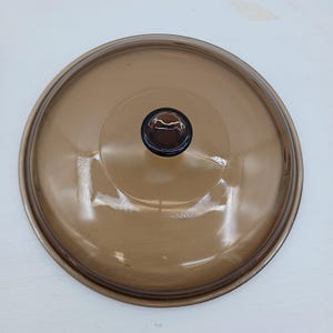 Könnte beinhalten: Ein runder, brauner Glasdeckel mit einem dunkelbraunen Knopf in der Mitte. Der Deckel ist transparent und ermöglicht einen Blick auf den Inhalt darunter. Der Deckel ist für Kochgeschirr bestimmt.