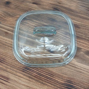 Op de afbeelding: Een helder, vierkant glazen deksel met afgeronde hoeken en een rechthoekige handgreep. Het deksel is ontworpen voor een kookpot of schaal. De handgreep is gemaakt van hetzelfde heldere glas.