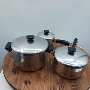 Può includere: Set di tre pentole in acciaio inossidabile con coperchi. Le pentole hanno manici e pomelli neri. Le pentole sono su una superficie rotonda di legno. Le pentole sono di diverse dimensioni.