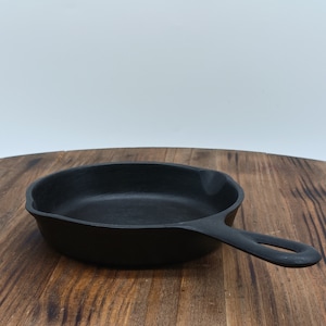 May include: A black cast iron skillet with a single handle. The skillet is sitting on a wooden surface.