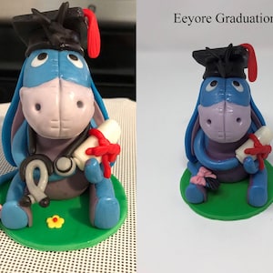 Puede incluir: Figura artesanal de Eeyore, el burro, con birrete y diploma atado con una cinta roja. La figura es principalmente azul, con un hocico rosa y una base verde. El texto "Eeyore Graduation" es visible.