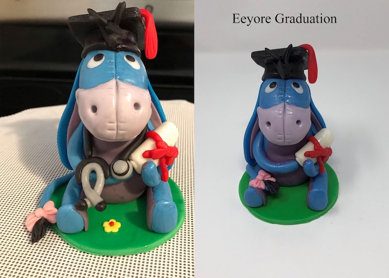 Puede incluir: Figura artesanal de Eeyore, el burro, con birrete y diploma atado con una cinta roja. La figura es principalmente azul, con un hocico rosa y una base verde. El texto "Eeyore Graduation" es visible.