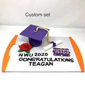Puede incluir: Un conjunto de graduaci&oacute;n personalizado con un birrete morado y borla dorada, un diploma enrollado, una rosa roja y un letrero que dice "Congrats Teagan". El conjunto est&aacute; sobre una tabla blanca con asas naranjas. El texto dice "NWU 2020 Congratulations Teagan".