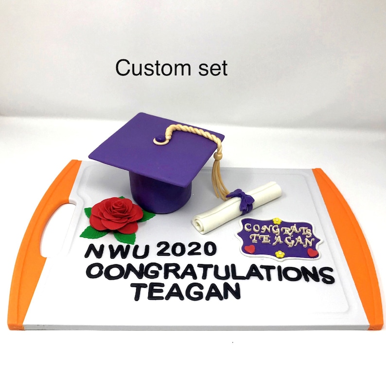 Puede incluir: Un conjunto de graduaci&oacute;n personalizado con un birrete morado y borla dorada, un diploma enrollado, una rosa roja y un letrero que dice "Congrats Teagan". El conjunto est&aacute; sobre una tabla blanca con asas naranjas. El texto dice "NWU 2020 Congratulations Teagan".