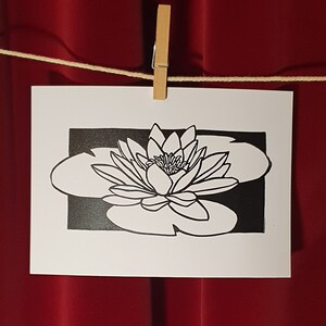 May include: Black and white line art print of a water lily with a single flower and leaves. The print is on white paper and is framed by a black border.
