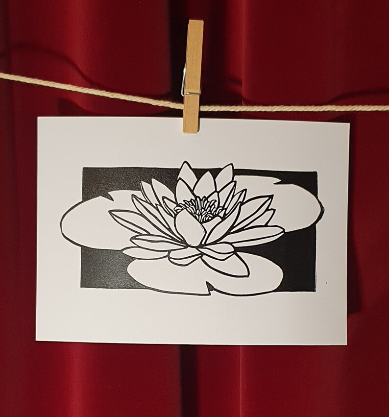 May include: Black and white line art print of a water lily with a single flower and leaves. The print is on white paper and is framed by a black border.