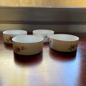May include: Set of four white ceramic bowls with a red rim and floral design. The bowls are on a wooden surface.