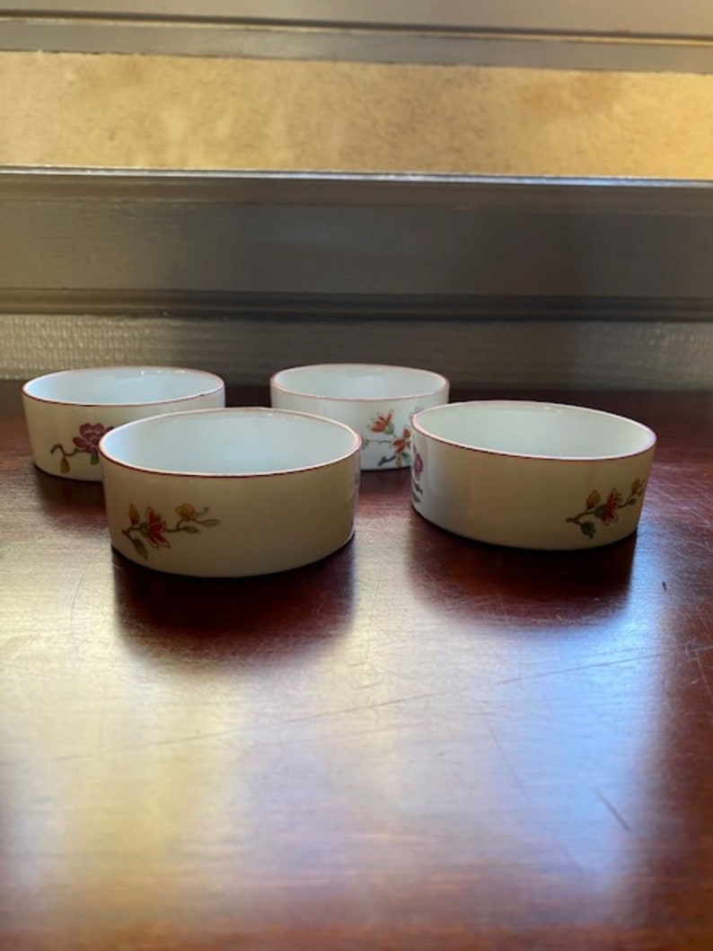 May include: Set of four white ceramic bowls with a red rim and floral design. The bowls are on a wooden surface.
