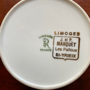 May include: Close-up of the bottom of a white porcelain plate with a gold and green logo that reads "LIMOGES J. et P. MARQUET Les Palloux St-YRIEIX FRANCE".