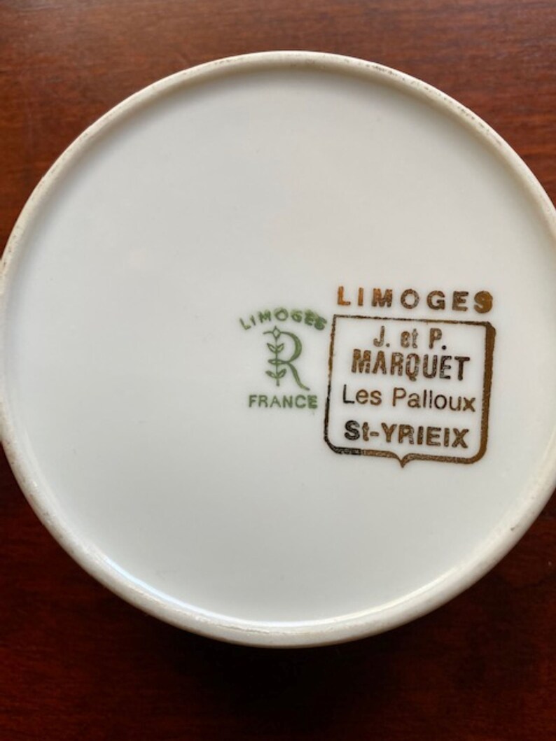 May include: Close-up of the bottom of a white porcelain plate with a gold and green logo that reads "LIMOGES J. et P. MARQUET Les Palloux St-YRIEIX FRANCE".