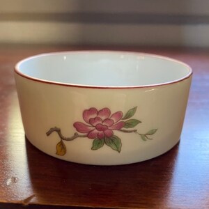 May include: A small white porcelain bowl with a red rim and a pink flower with green leaves and a yellow leaf on a branch design.