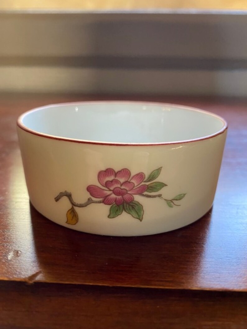 May include: A small white porcelain bowl with a red rim and a pink flower with green leaves and a yellow leaf on a branch design.