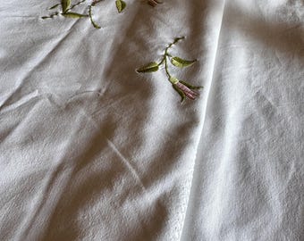 May include: White linen fabric with embroidered pink roses and green leaves. The embroidery is delicate and detailed, with a floral design. The fabric is folded, showing the texture and the embroidered edge.