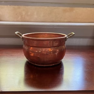 May include: A small copper pot with two silver handles. The pot has a rounded bottom and a slightly flared top. The pot is sitting on a wooden surface.