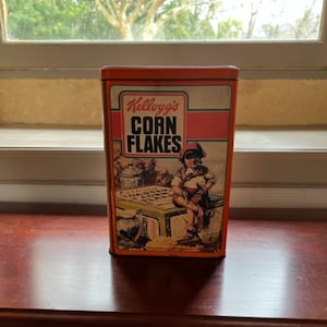 Op de afbeelding: Een vintage Kellogg's Corn Flakes-graanblik met een oranje achtergrond en een jongen in een rode hoed die op een doos zit. Het blik heeft een groen-witte streep rond de bovenkant en de woorden "Kellogg's Corn Flakes" in rood en wit.