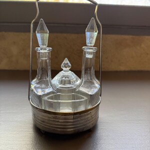 May include: A vintage condiment set with two glass bottles and a small dish, all held in a silver-toned metal frame. The bottles have diamond-shaped stoppers. The set is designed for serving condiments.