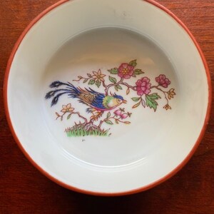May include: A small white porcelain dish with a red rim. The dish features a colorful bird with a long tail perched on a branch with pink flowers.