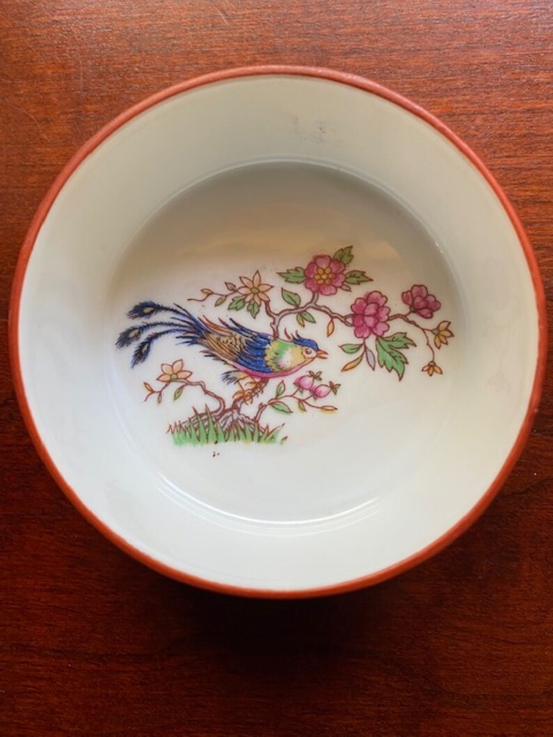 May include: A small white porcelain dish with a red rim. The dish features a colorful bird with a long tail perched on a branch with pink flowers.