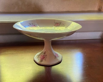 Puede incluir: Un tazón de pedestal de porcelana blanca con un diseño floral con rosas rosadas. El tazón tiene un borde festoneado y una base con pie.